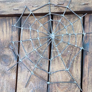 May include: A spider web sculpture crafted from dark grey metal wire. The design features a central hub with radiating spokes connected by concentric circular segments, all joined together with visible metallic welds at each intersection point.