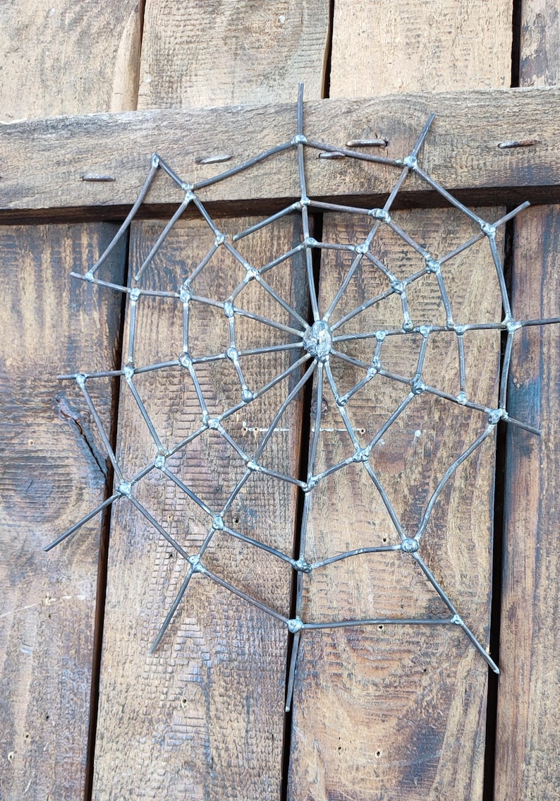 May include: A spider web sculpture crafted from dark grey metal wire. The design features a central hub with radiating spokes connected by concentric circular segments, all joined together with visible metallic welds at each intersection point.