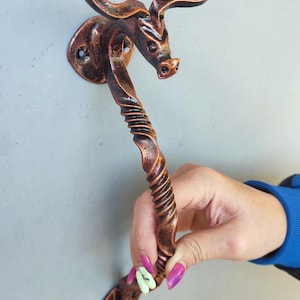 Könnte beinhalten: Ein bronzefarbener Türgriff mit einem Bullenkopf-Design. Der Griff hat einen gedrehten, strukturierten Griff und eine Montageplatte. Der Bullenkopf hat gebogene Hörner und detaillierte Gesichtszüge. Der Griff wird vor einem grauen Hintergrund gehalten.