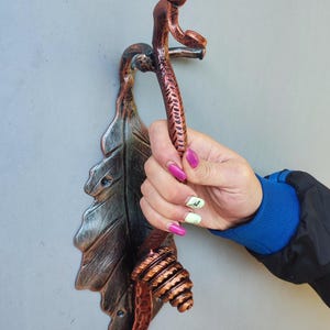 Könnte beinhalten: Ein dekorativer Türgriff aus kupferfarbenem Metall in Form eines Blattes und einer Weinrebe. Der Griff wird von einer Person gehalten. Der Griff hat ein Blattdesign und ein verdrehtes Weinreben-Design. Der Griff ist ca. 20 cm lang.