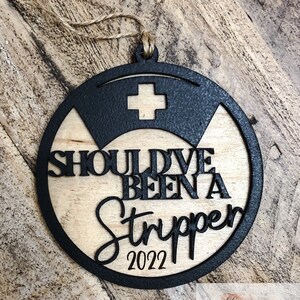 May include: A round wooden ornament with a black border and a white cross in the center. The ornament says "Should've Been a Stripper 2022".