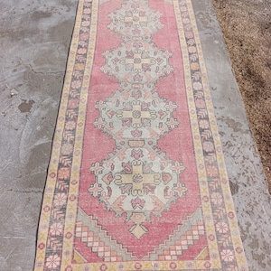 May include: A rectangular, vintage-style rug with a central pink field and intricate geometric patterns in cream, yellow, and gray. The rug has a border with repeating floral and geometric motifs. The rug is likely made of wool or a similar material.