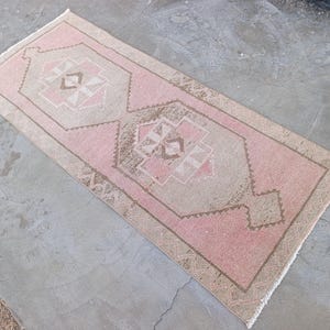 May include: A rectangular, vintage-style rug with a central geometric pattern in shades of pink, beige, and brown. The rug has a distressed look, with a border and fringe.