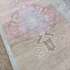 May include: A beige and pink vintage rug with a central floral design and geometric borders. The rug features a faded, distressed look, with the word "SUNA" visible in the design. The rug is rectangular and appears to be made of woven material.