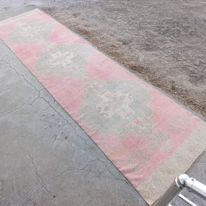May include: A long, rectangular rug with a pink and gray floral pattern. The rug is placed on a concrete surface. The rug's design features a central pink field with gray and white floral motifs.