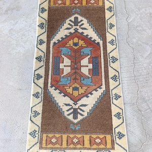 May include: A rectangular, hand-woven rug with a central brown field and a cream-colored border. The rug features a geometric design in shades of red, blue, yellow, and cream. The rug is likely made of wool and is suitable for a hallway or small space.
