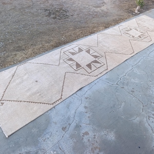 May include: A rectangular, beige runner rug with geometric brown patterns. The rug features repeating diamond and square designs along its length. The rug is placed on a concrete surface, showcasing its texture and design.