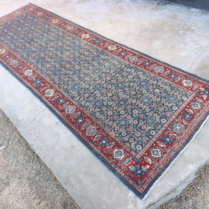 May include: A rectangular, patterned rug with a dark blue field and a red border. The rug features a repeating floral design in shades of blue, pink, and cream. The border has a detailed pattern with floral and geometric elements.