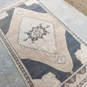 May include: Rectangular rug with a central diamond pattern in beige and dark gray. The rug has a border with floral and geometric designs. The rug is made of a woven material and is suitable for a living room or bedroom.