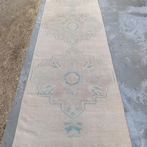 May include: A long, rectangular rug with a faded, vintage appearance. The rug features a central design with repeating geometric patterns in shades of beige, brown, and teal. The rug is likely made of woven material and is suitable for a hallway or entryway.