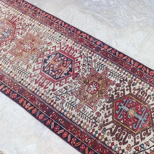 May include: A rectangular, vintage-style runner rug with a cream-colored background and intricate geometric patterns in red, blue, and gold. The rug features a central design with repeating motifs and a detailed border. The rug is likely made of wool.