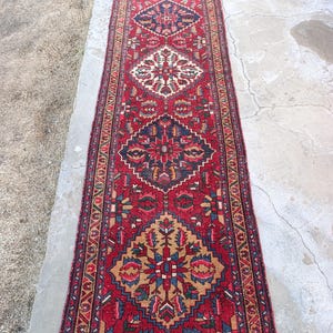 May include: A long, rectangular red rug with an intricate geometric pattern. The rug features a central design with diamond and floral motifs in shades of blue, beige, and gold. The rug's border is a darker red with a repeating pattern.