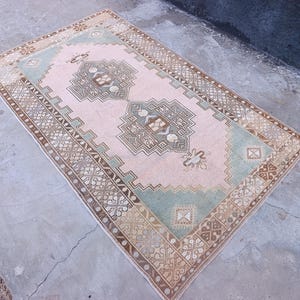 May include: Rectangular vintage rug with a central pink field, bordered by a brown and beige pattern. The rug features geometric designs in shades of green, brown, and beige, creating a traditional aesthetic.