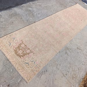 May include: A rectangular, vintage-style rug with a faded, distressed appearance. The rug features a central field of light beige with subtle pink hues. The border and a central medallion showcase floral patterns in shades of brown, pink, and blue.