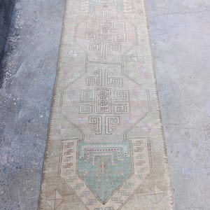 May include: A rectangular, vintage-style rug with a faded, distressed appearance. The rug features a central geometric design in shades of beige, pink, and teal. The rug's edges are slightly frayed, adding to its antique charm. The rug is suitable for a hallway or entryway.