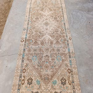 May include: A rectangular beige rug with an intricate, symmetrical pattern in shades of brown and teal. The rug features a central design with floral and geometric motifs, and a border with a repeating pattern. The rug has a fringe on the shorter ends.