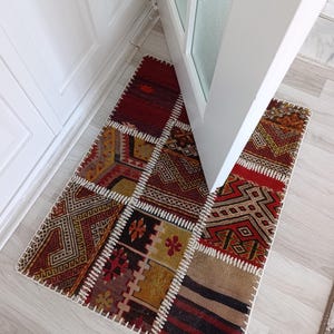 Puede incluir: Una alfombra rectangular de patchwork con una variedad de patrones geométricos en tonos rojos, marrones, dorados y beige. La alfombra está hecha de varias piezas rectangulares cosidas con costuras blancas. Se encuentra frente a una puerta blanca.