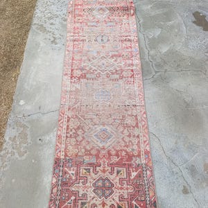 May include: A long, rectangular rug with an intricate, faded geometric pattern in shades of red, beige, and blue. The rug appears to be vintage or antique, with a distressed look. The rug is placed on a concrete surface.