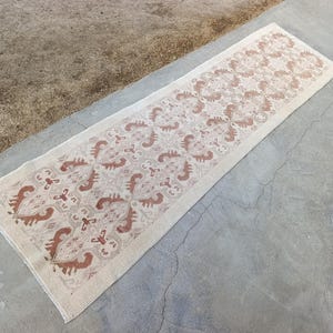 May include: A rectangular, cream-colored runner rug with a repeating pattern in shades of brown. The design features stylized floral and geometric shapes. The rug is placed on a concrete surface, showcasing its length and intricate details.