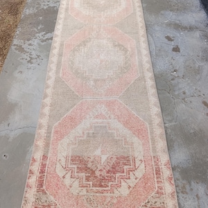 May include: A rectangular, distressed rug with a geometric pattern. The rug features a central design in shades of pink and red, framed by a beige border. The rug's design includes octagonal shapes and intricate details, suitable for a hallway or living space.
