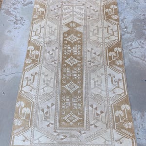 May include: Rectangular beige and brown rug with an intricate geometric pattern. The rug features a central panel with diamond and square motifs, framed by a border with stylized tree and floral designs. The rug's color palette is neutral, with variations in beige and brown.