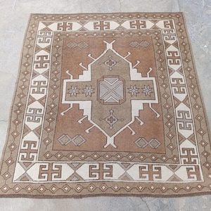 May include: A square, patterned rug with a central geometric design in shades of brown, beige, and white. The rug features a border with repeating geometric shapes. The rug is likely made of woven material and is suitable for home decor.