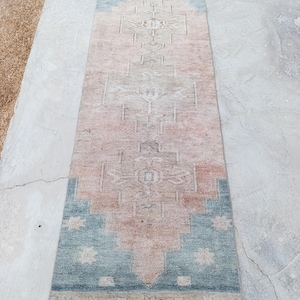 May include: A rectangular, vintage-style rug with a central geometric pattern. The rug features a color palette of light blue, peach, and beige. The design includes diamond and floral motifs, creating a traditional aesthetic. The rug is suitable for a hallway or entryway.