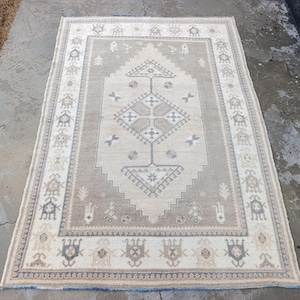 May include: A rectangular beige rug with a central geometric design in shades of brown and cream. The rug features a border with repeating patterns and a cream-colored background. The rug is placed on a concrete surface.