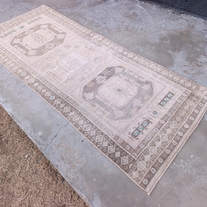 May include: A rectangular, vintage-style rug with a beige and brown color palette. The rug features intricate geometric patterns and a central medallion design. The border has a repeating diamond and floral motif. The rug is placed on a concrete surface.