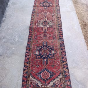 May include: A long, rectangular rug with a red background and intricate geometric patterns in navy blue, beige, and gold. The rug features a central medallion design and borders with repeating floral motifs. The rug is suitable for a hallway or entryway.