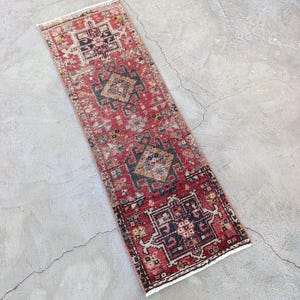 May include: A rectangular, distressed red rug with a geometric pattern in black, gold, and cream. The rug has a faded appearance and a fringed edge. The rug is placed on a concrete surface.