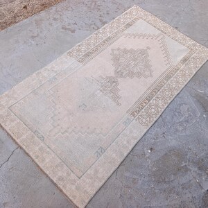 May include: A rectangular, vintage-style rug with a central medallion design and a detailed border. The rug features a muted color palette of beige, tan, and hints of blue-green, creating a soft, aged appearance. The rug is placed on a concrete surface.