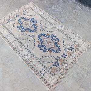May include: Rectangular beige rug with a central floral design in blue and brown. The rug features a detailed border with geometric patterns and a distressed appearance, suitable for home decor.