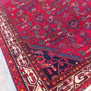 May include: A vibrant red oriental rug with intricate geometric patterns in blue, orange, and cream. The rug features a detailed border and a rich, textured surface, showcasing traditional craftsmanship.
