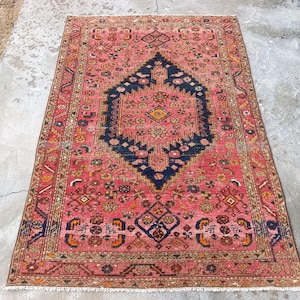 May include: A rectangular, vintage rug with a central navy blue medallion on a faded red background. The rug features intricate floral and geometric patterns in shades of orange, yellow, and brown. The rug has a fringed edge.
