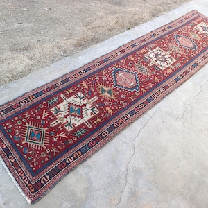 May include: A long, rectangular, red rug with intricate geometric patterns in white, blue, and beige. The rug features a dark blue border and a central design with repeating motifs. The rug is placed on a concrete surface.
