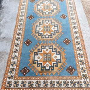May include: A rectangular, light blue rug with an intricate geometric pattern. The design features repeating star-like shapes in shades of orange, white, and brown. The border is a cream color with floral motifs. The rug is placed on a concrete surface.