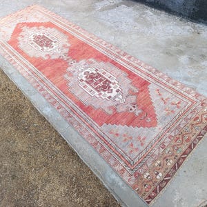 May include: A rectangular, distressed-look rug with a central medallion design. The rug features a color palette of coral, beige, and burgundy. Intricate border patterns frame the central design. The rug is placed on a concrete surface.