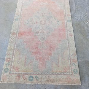 May include: A rectangular, vintage-style rug with a central design in shades of pink and gray. The rug features a detailed border with floral and geometric patterns in muted tones of pink, blue, and beige. The rug is placed on a concrete surface.
