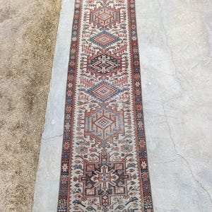 May include: A rectangular, patterned rug with a central design of geometric shapes in shades of red, blue, and brown on a cream background. The rug has a border with repeating patterns and is likely made of woven material.
