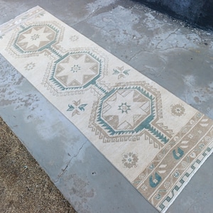 May include: A rectangular, cream-colored rug with geometric patterns in shades of beige and teal. The rug features a central design of repeating star-like shapes within octagons, bordered by a detailed edge. The rug is placed on a concrete surface.