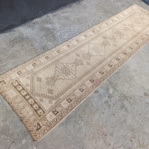 May include: A rectangular, beige-colored runner rug with an intricate, geometric pattern. The rug features a central design with repeating motifs and a border with floral and geometric elements. The rug is placed on a concrete surface.