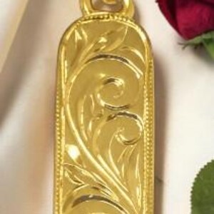 May include: A gold-colored pendant with an ornate, engraved floral design. The rectangular pendant has rounded edges and a loop at the top for a chain. The pendant is highly reflective and appears to be made of metal. The background includes red roses.