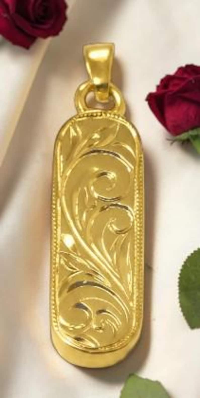 May include: A gold-colored pendant with an ornate, engraved floral design. The rectangular pendant has rounded edges and a loop at the top for a chain. The pendant is highly reflective and appears to be made of metal. The background includes red roses.