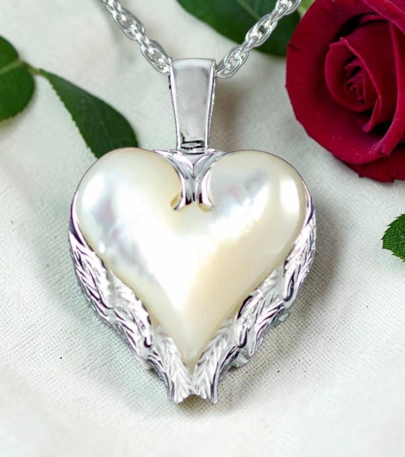 May include: A heart-shaped pendant necklace with a pearlescent center and silver-toned wing accents. The pendant hangs from a silver chain. The necklace is a piece of jewelry.