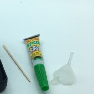 May include: A green tube of glue with a white applicator tip, a small plastic funnel, and a wooden stick. The glue tube has the text "Super Glue" printed on it.
