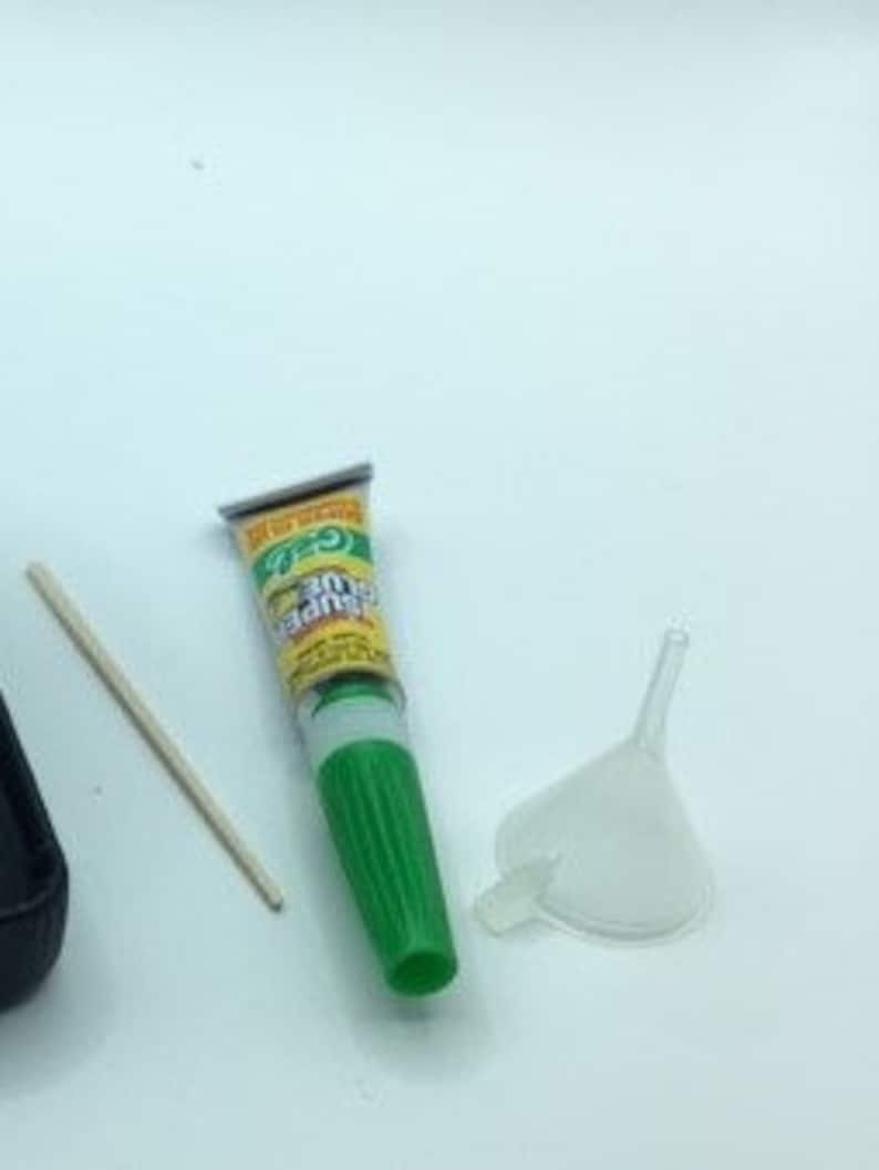 May include: A green tube of glue with a white applicator tip, a small plastic funnel, and a wooden stick. The glue tube has the text "Super Glue" printed on it.