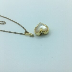 May include: A gold-colored necklace with a heart-shaped pendant. The pendant has a pearlescent, off-white center and a decorative gold frame. The necklace chain is also gold-colored and lies on a white surface.