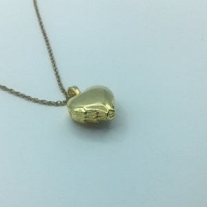 May include: A gold-colored heart-shaped pendant necklace. The pendant has a textured surface and is suspended from a delicate chain. The necklace is displayed against a white background.