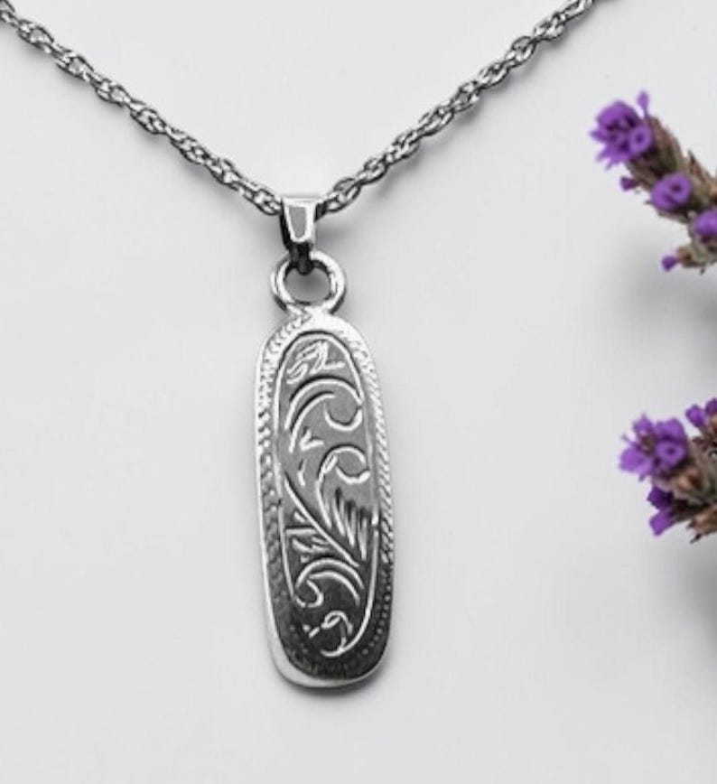 May include: A silver pendant necklace with an oval-shaped pendant. The pendant features an engraved floral design and hangs from a delicate chain. The background is white with a sprig of purple flowers.